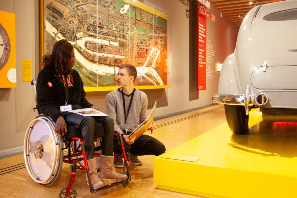eine Frau im Rollstuhl und ein mann sitzen vor einem hafenmodell im Museum