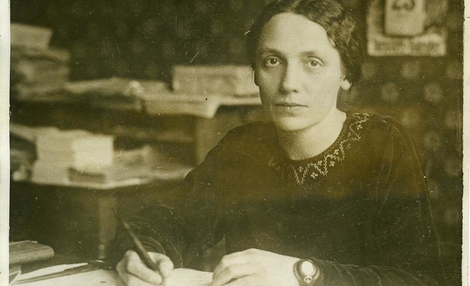 schwarz-weiss-Foto: eine Dame sitzt am Schreibtisch, sie hat einen Stift in der Hand, vor ihr liegt Papier