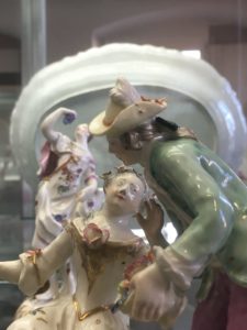 Detail zweier, einander zugewandter Porzellanfiguren. Eine Frau sitzt und blick zu einem Mann der Blumen in einem Korb hält