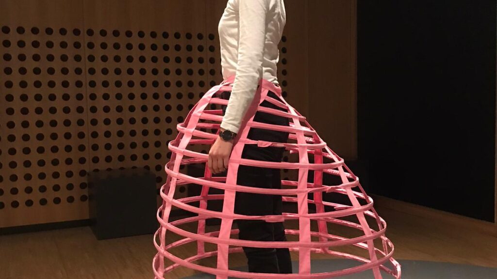 eine Frau steht mit einer Krinolinenform - einen Unterrockgestehl aus Kunststoff im Ausstellungsraum, sie ist von der Seite zu sehen - nach hinten ragt das gestell noch heraus
