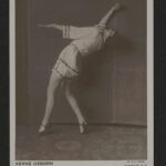 eine schwarzweiß-Fotografie mit einer weissgekleideten Tänzerin mit kurzen Hosen, die sich nach hinten lehnt und dabei den rechten Arm hochstreckt