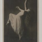 Schwarzweiß-Fotografie mit einer Tänzerin im weißen Kleid. Sie beugt sich nach hinten, und hat das linke Knie angebeugt und den rechten Arm nach oben ausgestreckt.