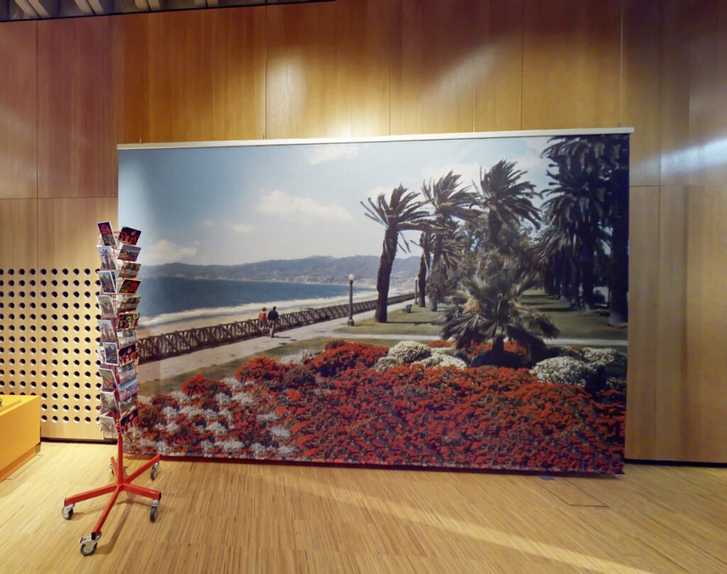 ein riesiges Transparent mit geranien und Palmen am meer, davor ein Postkartenhalter