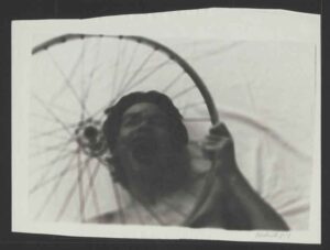 Eine etwa unscharfe Fotografie zeigt einen schreienden Mann, der ein Rad in die Hand hält. Es handelt sich um den japanischen Tänzer Mutsuo Hirano.