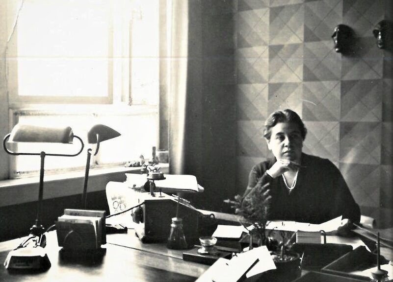 Die S/w-Fotografie zeigt eine Frau am Schreibtisch. Vor ihr auf dem Tisch steht eine große Schreibmaschine