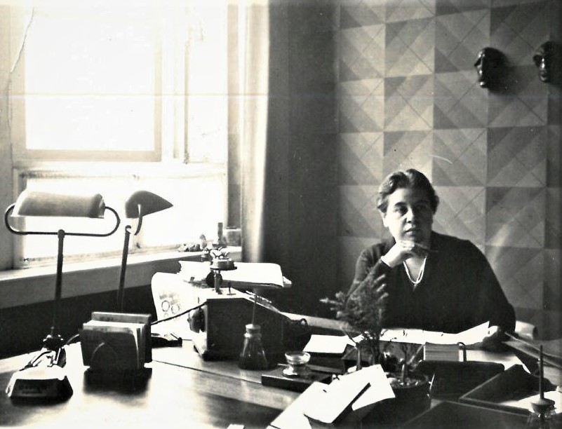 Die S/w-Fotografie zeigt eine Frau am Schreibtisch. Vor ihr auf dem Tisch steht eine große Schreibmaschine