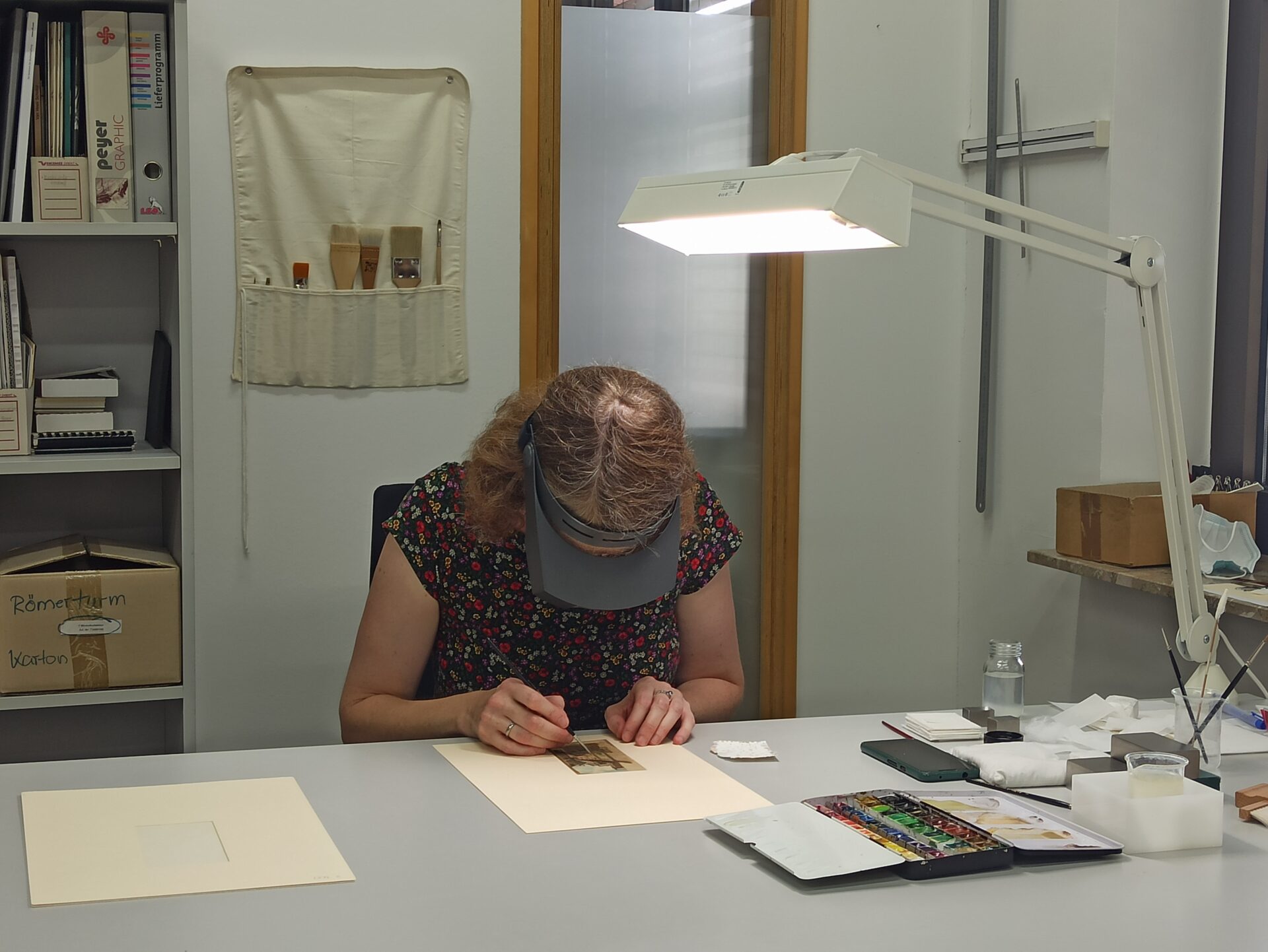 Die Papierrestauratorin sitzt an einem Tisch in ihrer Werkstatt. Sie untersucht und retuschiert eine Aquarelle mit einer speziellen Brille