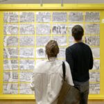 Zwei Ausstellungsbesucher:innen stehen vor einem Ausstellungsbeitrag und schauen ihn an. Dieser besteht aus 42 schwarz-weiß Zeichnungen, die zusammen ein großes fiktives Bild von Frankfurt zeichnen.
