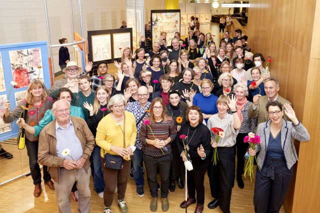 Gruppenfoto von etwa 50 Personen mit Blumen in der Hand. Sie stehen in der Ausstellung.