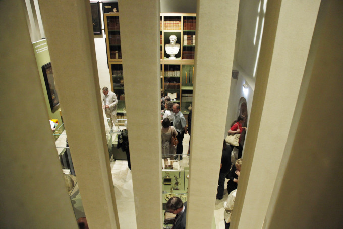 Im Vordergrund des Bildes sind Stelen zu sehen. Der Blick hindurch zeigt einen Ausstellungsraum im Sammlermuseum. Einige Objekte sind in Vitrinen ausgestellt. An der Rückwand ist eine große Bücherwand und eine Büste zu sehen. Zwischen den Vitrinen stehen einige Personen.