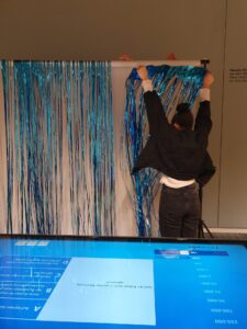 Eine der Kuratorinnen steht mit dem Rücken zur Kamera und befestigt blaues Lametta an einer weißen Wand. Im Vordergrund ist ein Bildschirm mit eienm Quiz zu sehen.