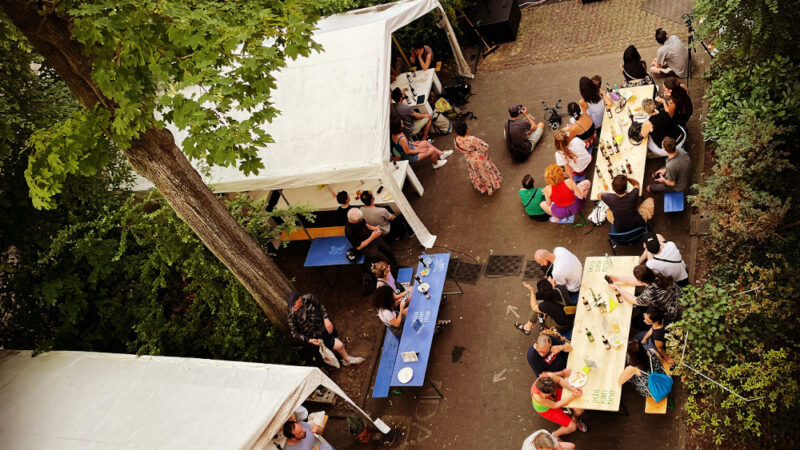 Der Hinterhof der ada_kantine ist von oben fotografiert. In und zwischen drei großen Zelten stehen Biertischgarnituren. An den Tischen sitzen viele Menschen.