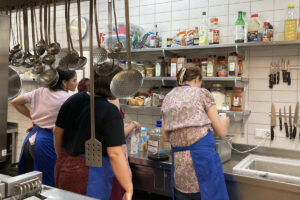 Vier Menschen stehen in einer industriellen Küche und arbeiten an einem Gericht. Im Hintergrund ist ein Gewürzregal zu sehen.
