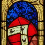 Eine Wappenscheibe aus Glas zeigt ein rotes Wappen mit einem weißen Band. Darauf sind drei Hühner zu sehen. Der Hintergrund besteht aus blauen und goldenen Ornamenten.