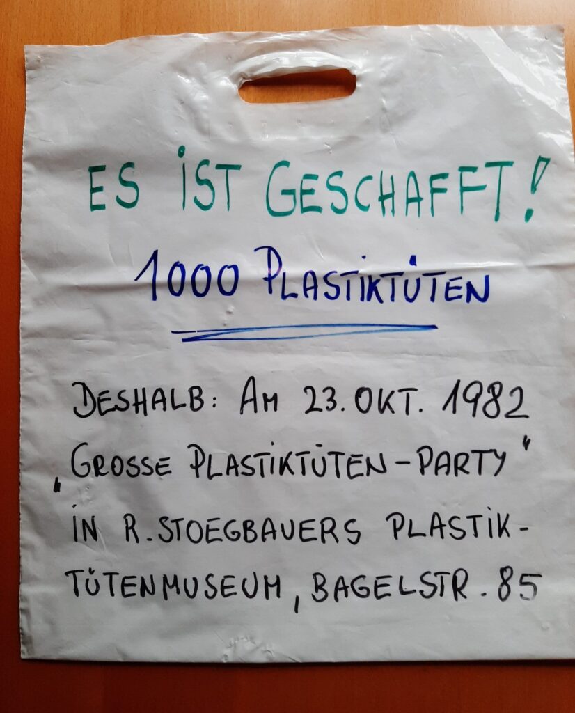 Auf einer weißen Plastiktüte steht in Druckschrift mit einem Filzstift geschrieben:Es ist geschafft! 1000 Plastiktüten Deshalb: am 23. Okt. 1982 "Grosse Plastiktüten-Party" in R. Stoegbauers Plastiktütenmuseum, Bagelstr. 85