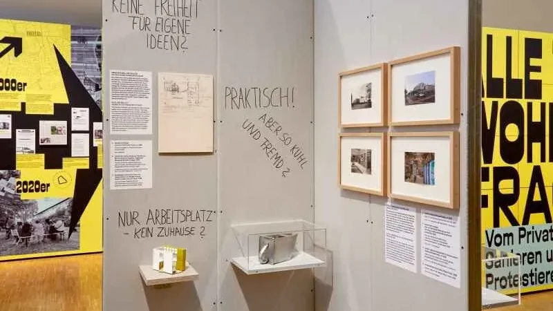 Blick auf eine Wand in einer Ausstellung mit gerahmten Bildern und kleinen Vitrinen