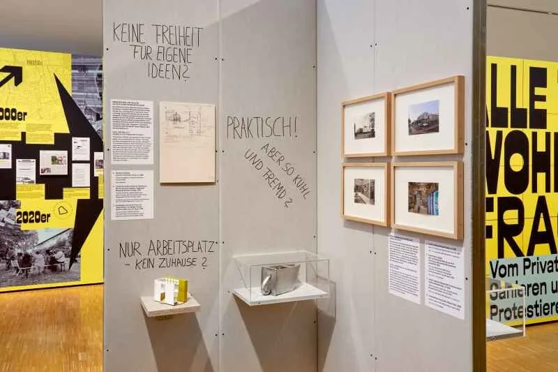 Blick auf eine Wand in einer Ausstellung mit gerahmten Bildern und kleinen Vitrinen